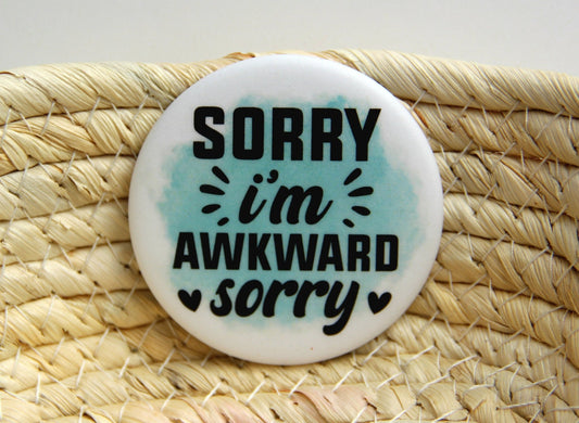 A round white refrigerator magnet with a black quote. The saying reads Sorry I'm awkward sorry. A watercolor style splash of teal is behind the quote.