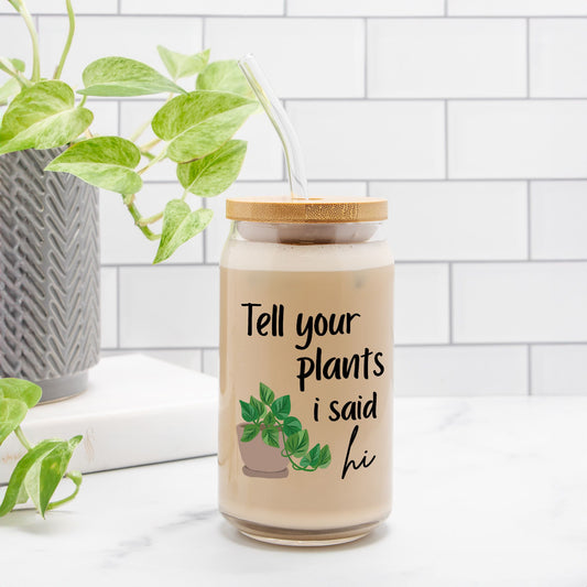 A glass with iced coffee with a quote that reads Tell your plants i said hi. There is a green houseplant with a brown pot.